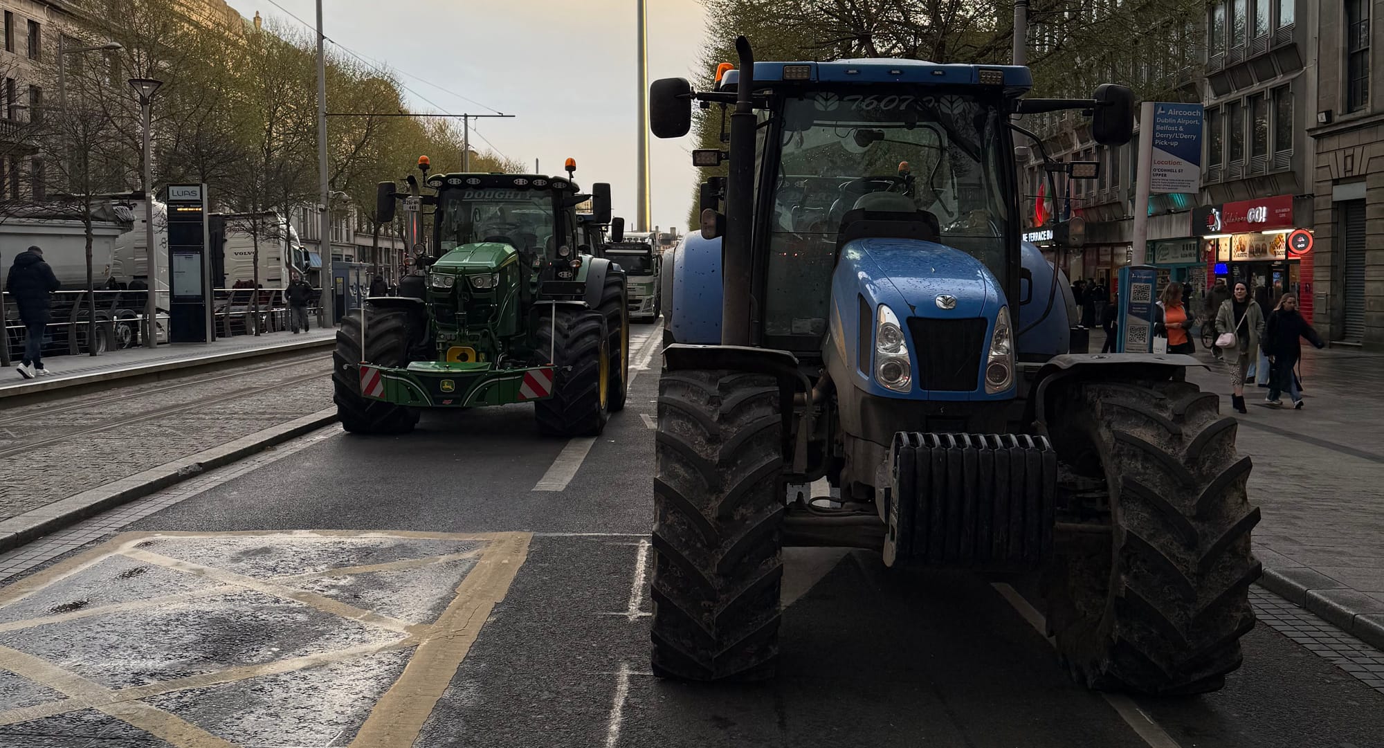 A green tractor cab and a blue tractor cab are facing the camera. They are parked on a city street. There is a tram line on the left hand side and a footpath on the other.