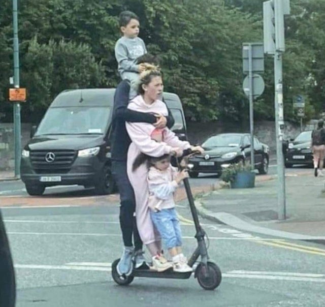 A man and a woman on an e-scooter. The man has a toddler on this shoulders and there is a small child standing in front of the woman on the scooter.