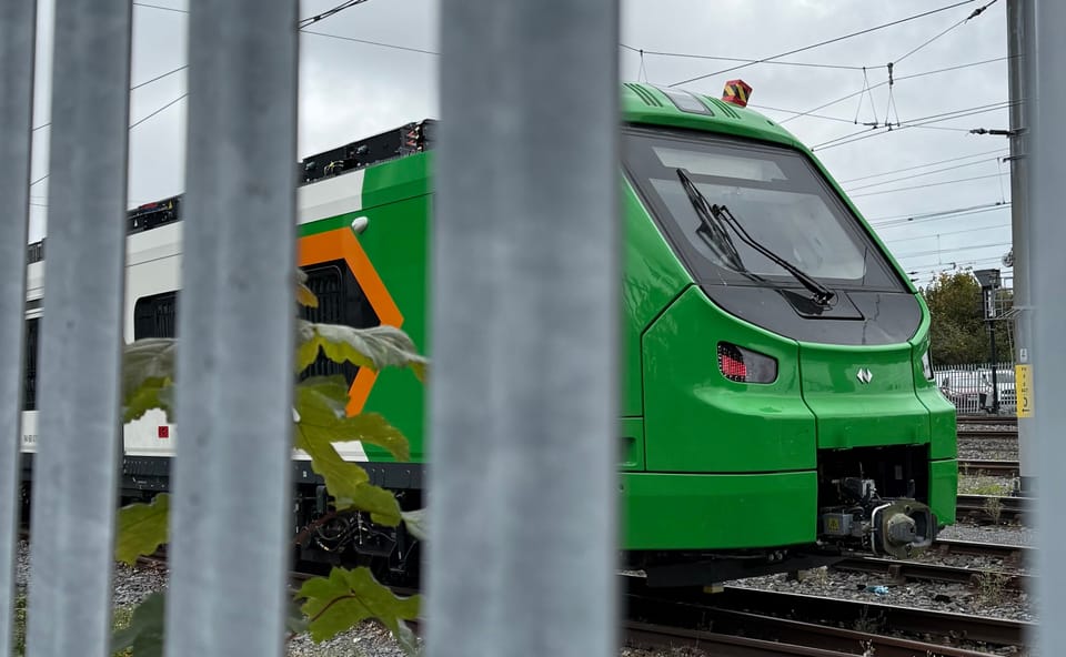 A green train with flashes of orange and white livery viewed through a railing